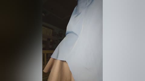 A video of a person standing in a dimly lit room, wearing a white shirt and beige pants, with a blurred background featuring wooden furniture and a bookshelf.