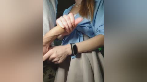 Video of a woman in a blue shirt and beige skirt, holding a man's erect penis. She has light skin, a black wristwatch, and her hair is tied back. Background is blurred.