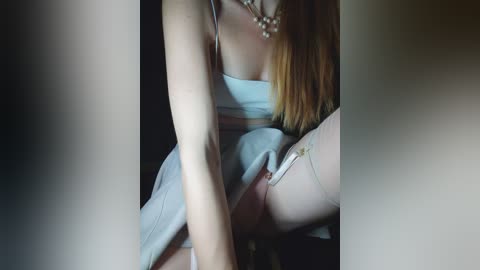 Video of a slender, fair-skinned woman with long, straight, blonde hair, wearing a light blue dress and white stockings, seated in a dimly lit room.