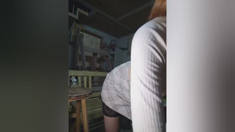 Video of a woman with light skin, wearing a plaid skirt and black fishnet stockings, leaning over a wooden chair in a dimly lit room.