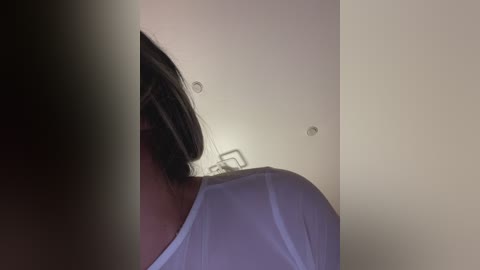 Media: Video of a woman's bare shoulder and back, with long dark hair, wearing a white shirt, in a dimly lit room with beige walls and two small ceiling lights.