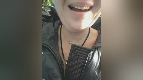 Video of a smiling, light-skinned person wearing a black puffy jacket and a black necklace, outdoors with blurred green foliage in the background.