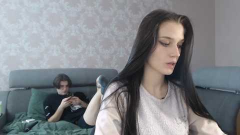 Media: A video of a young woman with long black hair, wearing a white sweater, drying her hair. Behind her, a man in a black shirt plays a video game.