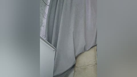 A video of a partially draped grey curtain covering a beige wall and part of a white bathtub, with a visible white pipe and a textured, off-white floor. The lighting is soft and diffused.