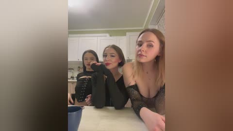 Video of three young women, two with long hair, one with short hair, in a kitchen, wearing black lingerie, leaning on a counter.