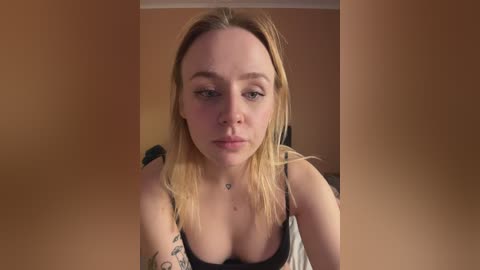 Video of a blonde woman with fair skin and a medium build, wearing a black tank top, sitting in a beige room with minimal background details.