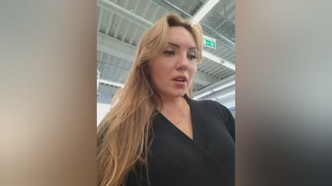 Video of a blonde woman with long hair, wearing a black V-neck sweater, standing in an airport with a metallic ceiling and visible signage.