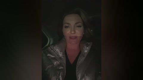 Video of a Caucasian woman with light brown hair, wearing a silver puffy jacket, sitting in a car at night. She has a solemn expression.
