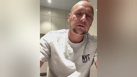 Media: Video of a bald, muscular white man with a beard wearing a gray t-shirt, standing in a modern kitchen with white cabinets and recessed lighting.