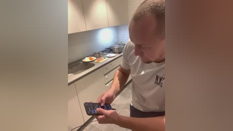 Media: Video of a bald, fit man in a white t-shirt using a smartphone while cooking in a modern kitchen with beige cabinets and a stainless steel sink.