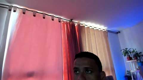 Video of a young Black man with short hair, wearing a gray shirt, looking toward a partially open window with a pink and beige curtain.
