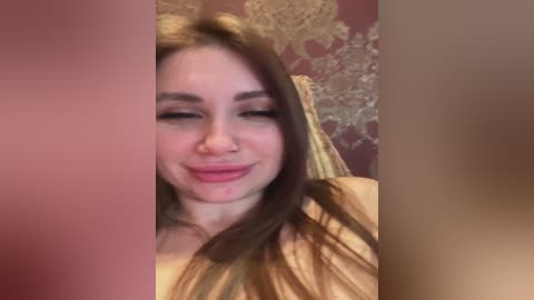 Media: Video of a young woman with fair skin, long brown hair, and a nose piercing, smiling while wearing a golden crown. The background features a patterned red wall.