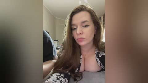 Media: A video of a young Caucasian woman with long brown hair, wearing a black floral dress, lying on a bed, making a pouty face, with blurred background.