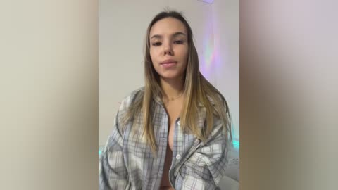 Video of a young woman with light brown skin and long, straight, blonde hair, wearing a plaid shirt, smiling, standing against a plain beige wall with soft, multicolored LED lights in the background.