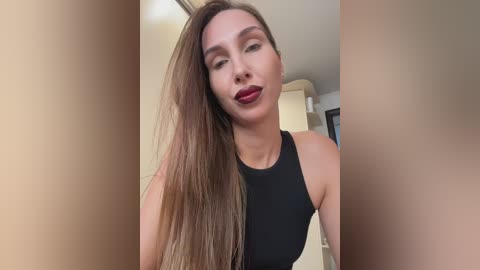Video of a light-skinned woman with long, straight brown hair, wearing a black sleeveless top, standing indoors with beige walls and a white shelf in the background.