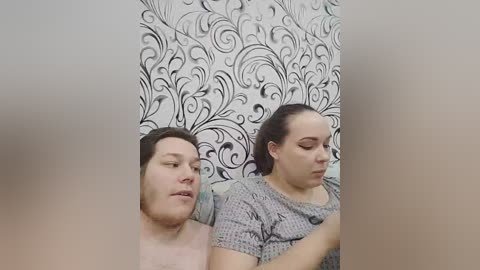 Video of two overweight people with light skin, seated side by side. The man on the left has short dark hair and a beard, wearing a pink shirt. The woman on the right has dark hair pulled back, wearing a gray patterned shirt. They appear in a room with black and white floral wallpaper.