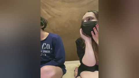 Video: Two women, one with short hair, wearing a navy sweatshirt, and the other with long hair, black mask, and black top, sit against a brown backdrop.