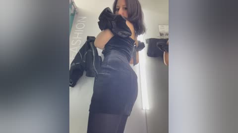 Video of a woman with medium skin tone and shoulder-length black hair, wearing a tight, black satin dress with puffed sleeves, standing in a dimly lit room.