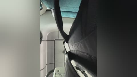 Media: Video of an airplane seat viewed from below, showing a person's legs in dark pants, a turquoise fabric seat, and a white armrest. The image captures the texture of the fabric and the metal armrest.