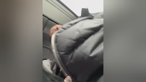 Media: A video captures a close-up of a person's upper body inside a car, with their hands gripping a gray, puffy jacket. The background features a car's interior with a visible window and light-colored ceiling.