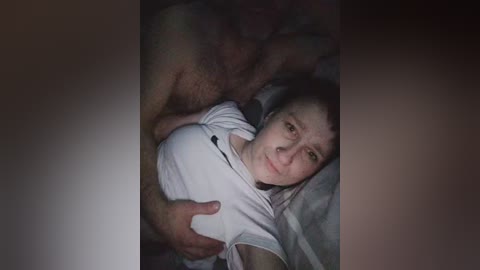 Video of a topless man embracing a white woman with fair skin, wearing a white t-shirt, lying on a bed with gray sheets, dim lighting.