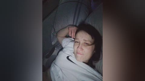 Video of a young woman with fair skin, brown hair, and light-colored clothes lying on a bed with gray pillows, looking tired and holding her head.