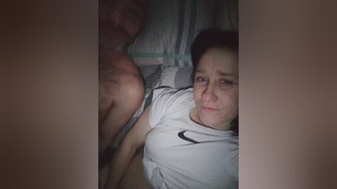 A video captures a close-up of a woman lying on a bed, wearing a white Nike t-shirt, with a man's bare chest and face partially visible in the background.