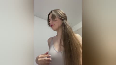 A video of a Caucasian woman with long, straight brown hair, wearing a white tank top, standing in a corner of a white-walled room. She has fair skin and red lipstick.