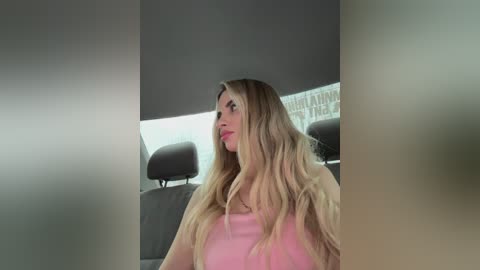 Media: Video of a blonde woman in a light pink top, sitting in a car with gray seats and headrests, looking to the side.