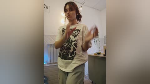 Media: Video of a young woman with medium-length brown hair, wearing a white T-shirt with a graphic design, gray sweatpants, and a casual expression. She's in a small, dimly-lit room with white walls and a cluttered background.