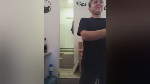 Media: A video of a young man with light skin and short brown hair, wearing a black t-shirt and jeans, standing in a bathroom with a wall-mounted water dispenser and a tiled shower in the background.