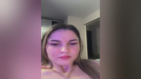 Video of a young woman with light skin, straight brown hair, and full eyebrows, wearing a gold necklace, taken indoors with blurred background.