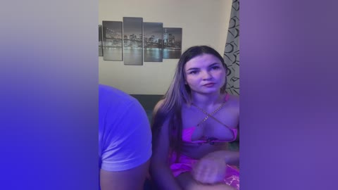 A video captures a young woman with long, straight brown hair, wearing a pink bikini, seated on a couch. The background features cityscape artwork and a purple curtain.