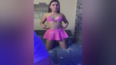Video of a young woman with fair skin and long brown hair, wearing a shiny pink bikini top and mini skirt, kneeling on a bed in a dimly lit room with blue lighting.