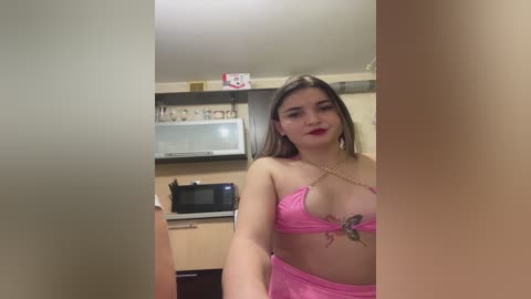 A video of a young woman with fair skin, straight brown hair, and a slim figure, wearing a revealing pink lace lingerie set and a gold chain necklace, posing seductively in a modern kitchen with wooden cabinets and a microwave.