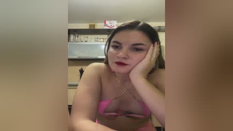 Video of a young woman with fair skin and dark hair, wearing a pink bikini with a gold chain, resting her chin on her hand in a kitchen with beige cabinets and a red sign.