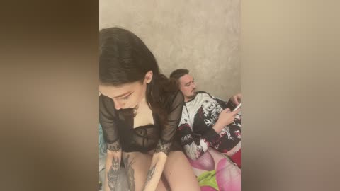 Media: Video of a tattooed woman with long dark hair wearing a sheer black top, sitting on a man with a short beard in a floral kimono, against a beige textured wall.
