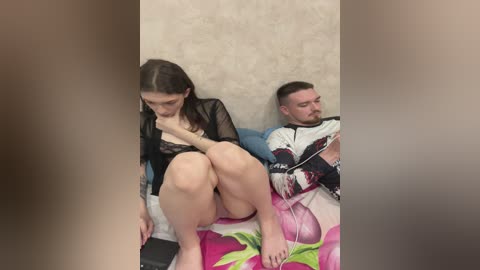 Media: A video of a young woman with fair skin and dark hair, sitting on a bed, wearing a black lace bra, looking down. Beside her, a man with a short beard, wearing a white shirt with floral patterns, listens to headphones.