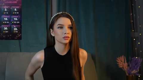 Media: Video of a young, slender woman with long brown hair wearing a black sleeveless top, seated indoors. Background features a teal curtain, a weather forecast screen, and a decorative purple plant.