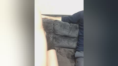 A video shows a person wearing a dark blue jacket, partially obscured, lifting a large, soot-covered, black leather couch against a plain white wall. The scene appears to be indoors, possibly in a garage or storage area.