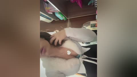 A blurry video shows a woman with fair skin, wearing a white fur-trimmed coat and black pants, adjusting her hair in a messy room.