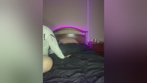 Media: A video of a messy bedroom with a person in white pajamas and a black shirt, lying on a bed with a disheveled black blanket. Purple LED lights adorn the wall behind a wooden headboard.