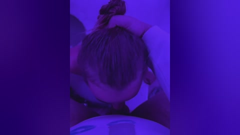 Media: Video of a woman with brown hair, wearing a white shirt, leaning over a table, with purple lighting creating a moody atmosphere.