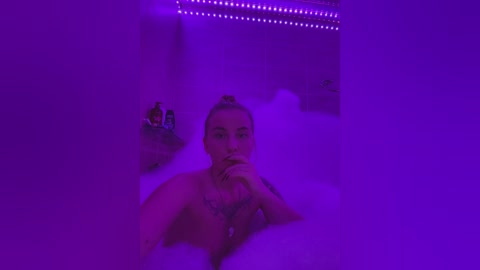 Media: Video of a topless, fair-skinned woman with blonde hair in a bun, gazing pensively, partially obscured by steam. Purple lighting casts a moody, intimate ambiance in a modern, dimly lit bathroom.