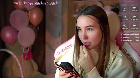 Media: A video shows a young Asian woman with long black hair, wearing a beige jacket, holding a smartphone. She's indoors, surrounded by pink balloons and a pink and white gaming chair.