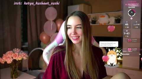 Media: Video of a smiling young woman with long straight hair, wearing a deep red shirt, surrounded by pink balloons in a cozy room.