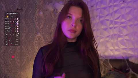 Video of a young woman with long, straight brown hair, wearing a black sheer top, against a textured, dimly lit background.