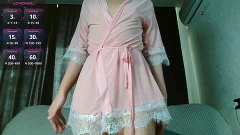Video of a slender, light-skinned person in a pink, short robe with white lace trim, standing in a dimly lit room with a bed, air conditioner, and curtains.
