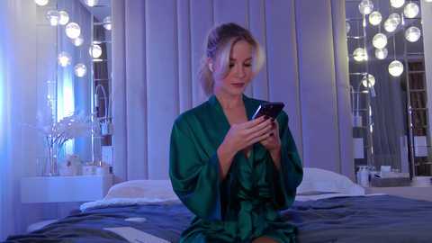 Video of a blonde woman in a green satin robe, sitting on a bed, engrossed in her smartphone, in a chic, dimly lit bedroom with mirrored walls and spherical lamps.