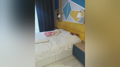 Media: A video of a modern, cozy bedroom with a white bed, yellow upholstered headboard, and colorful geometric-patterned wall. A stuffed toy duck rests on the bed, and a nightstand with a lamp is nearby.